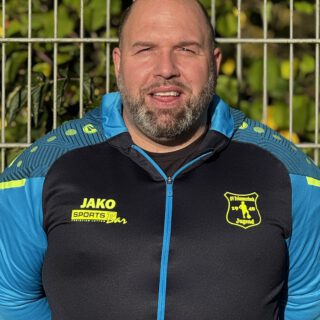 Dennis in blau-schwarzem Trainingsjacke mit gelben Logos von JAKO Sports Bar und SV Frömmersbach Jugend, vor einem metáallenen Zaun, nahaufnahme Gesicht