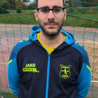 Mann mit Brille trägt eine Sportjacke mit Vereinslogo und steht vor einem Fußballfeld.