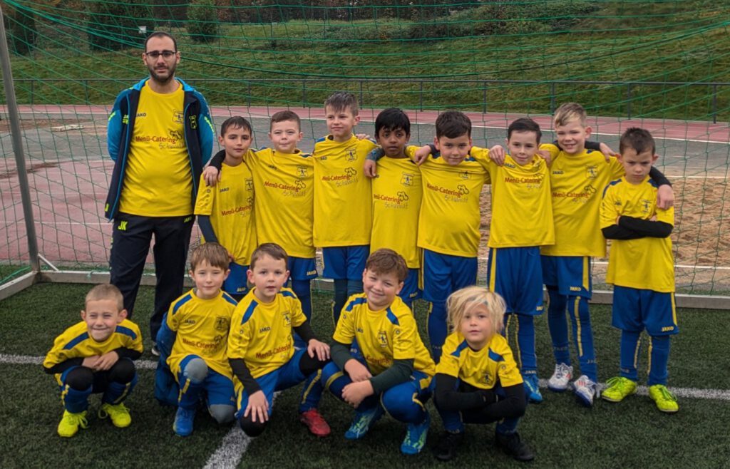 Eine U8 Fußballmannschaft mit gelben Trikots steht auf einem Sportplatz. Trainer und Spieler posieren für das Foto.