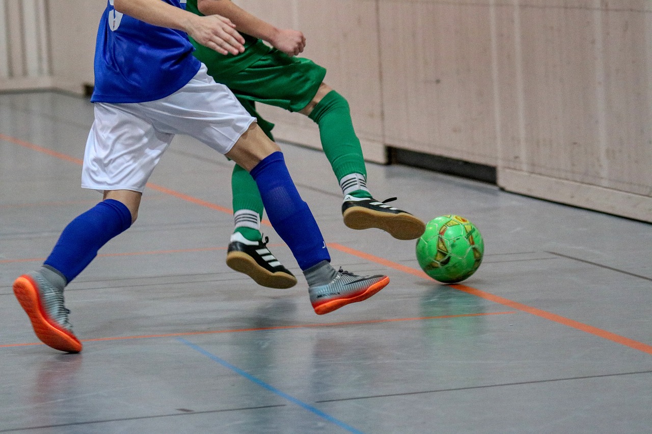 Zwei Spieler in blauen und grünen Trikots, die einen Fußball in einer Halle spielen.