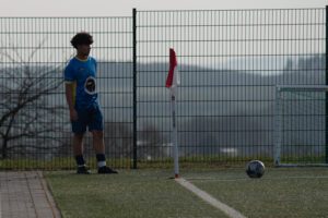 Junger Fußballspieler im blauen Trikot steht am Spielfeldrand eines Kunstrasenplatzes neben der Eckfahne, vor einem hohen Zaun; Ball liegt nahe dem Tor.