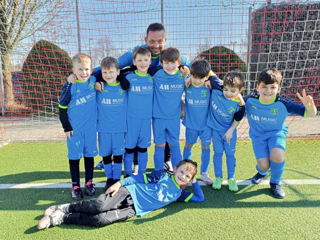 Gruppe junger Fußballspieler in blauen Trikots posiert lachend vor dem Tor, Trainer im Hintergrund, sonniger Tag; Verein SV Frömmersbach sichtbar in der Szene