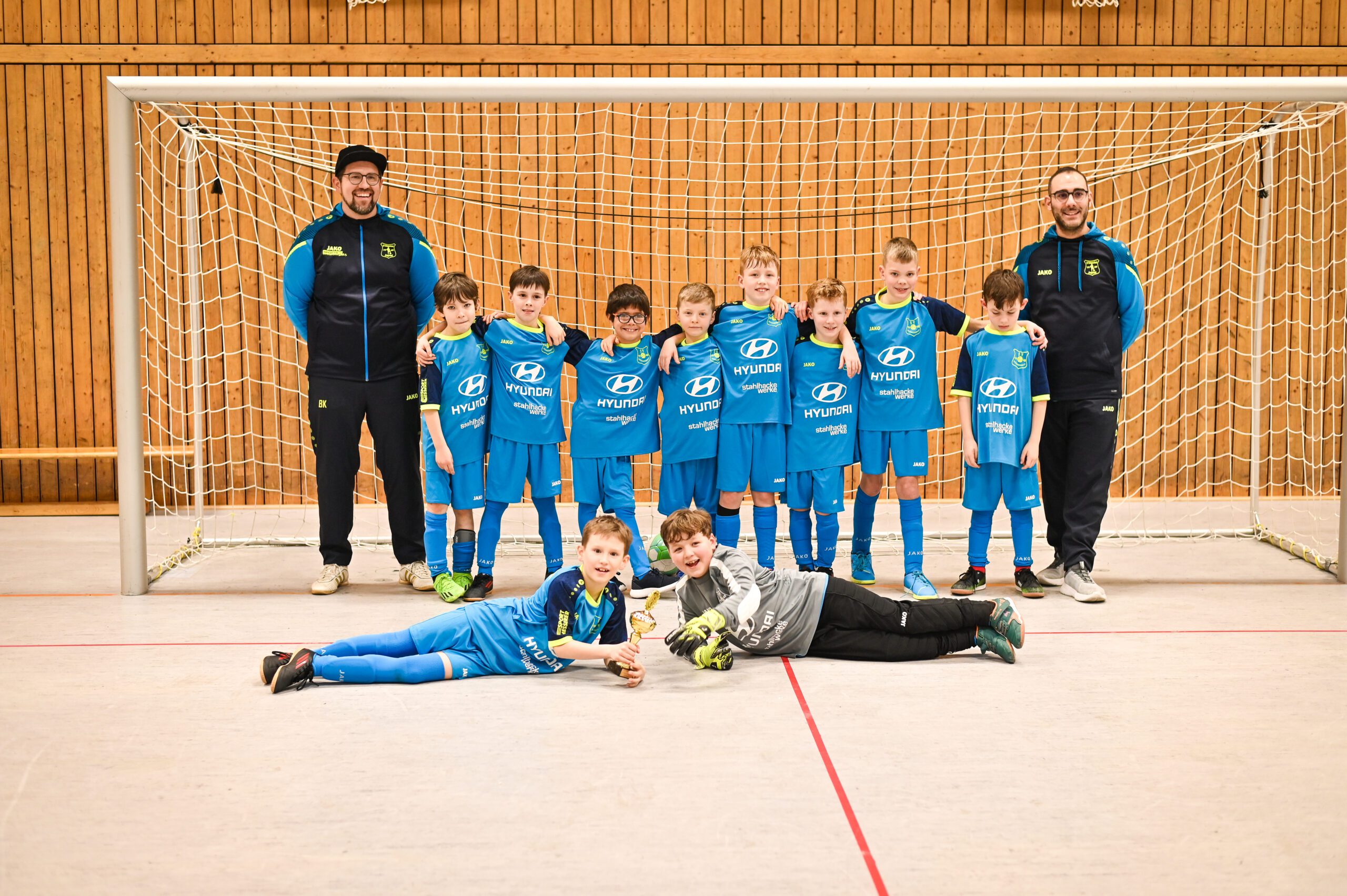 Junges Fußballteam in blauen Trikots posiert vor einem Tor in einer Halle; zwei Trainer stehen seitlich, alle Jungen schmunzeln und tragen Hyundai-Logo-Trikots.