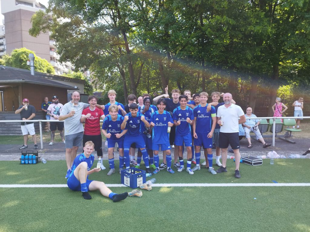 Die A-Junioren des SV Frömmersbach feiern in blauen Trikots mit Trainern und Fans den Aufstieg, während Becher und Kisten am Spielfeldrand stehen.